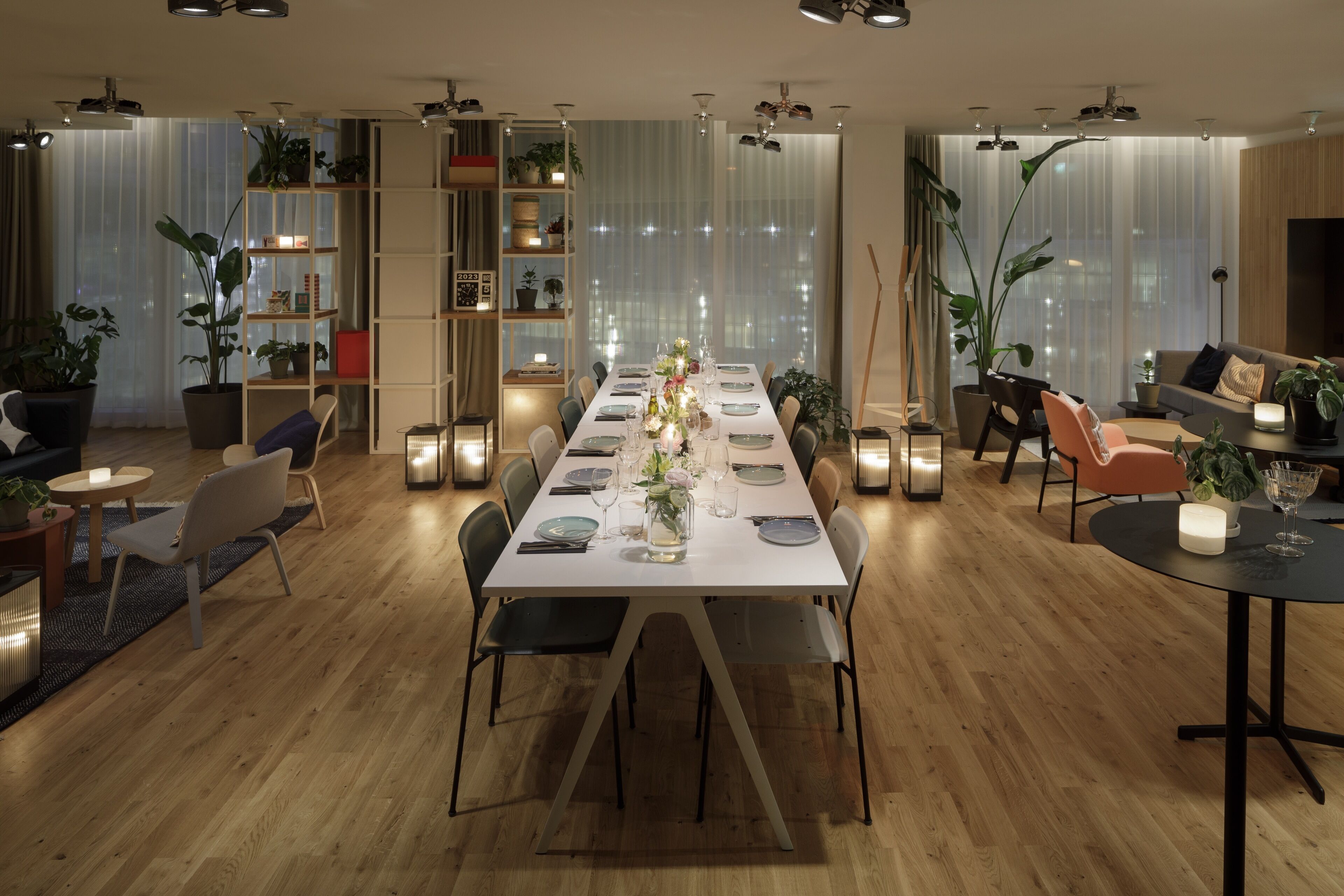 A modern dining area with a long table set for six, surrounded by chairs. There are shelves with plants and books on the walls, and a sofa and armchairs in the background.