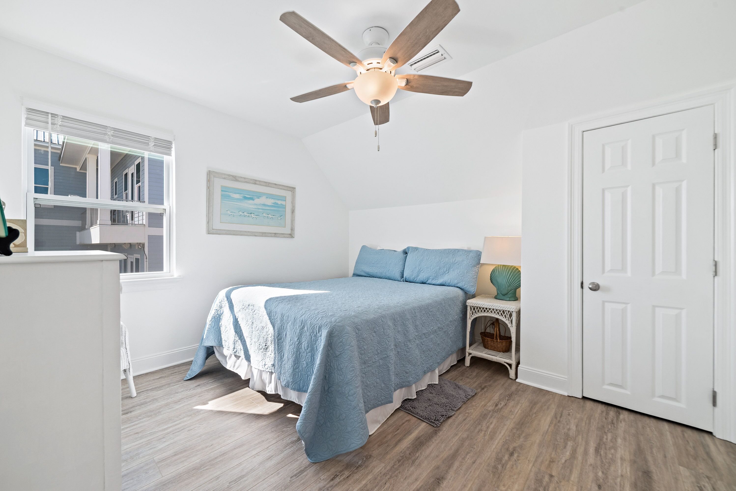 A bedroom with a bed, a ceiling fan, a window, a door, and a nightstand with a lamp.