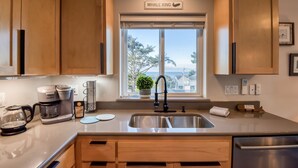 A kitchen with wooden cabinets, a stainless steel sink, a coffee maker, and a window with a view of the outdoors. - Pacific Peeks 2 - Oceanview Pet Friendly Retreat with EV Charging (fee) (Lincoln City)
