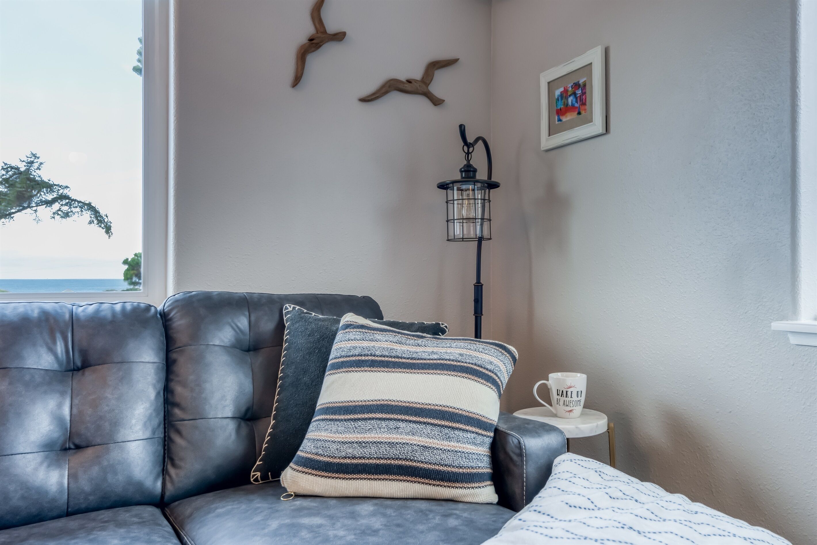 A room with a leather sofa, striped pillow, a small table with a mug, a wall-mounted lamp, and a framed picture.