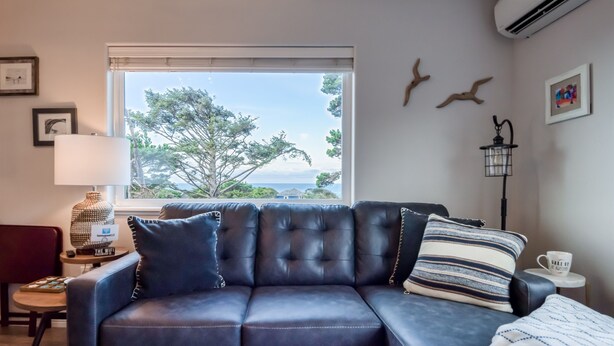 A dark blue leather sofa with multiple cushions, a lamp, a small table with a cup, and a view of trees and a house through a window. - Pacific Peeks 2 - Oceanview Pet Friendly Retreat with EV Charging (fee) (Lincoln City)