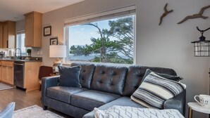 A modern living room with a dark leather sofa, a patterned throw pillow, and a view of the sea through a large window. - Pacific Peeks 2 - Oceanview Pet Friendly Retreat with EV Charging (fee) (Lincoln City)