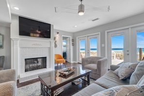 A living room with a fireplace, a glass coffee table, and a sofa. There is a television mounted above the fireplace and a door leading to a balcony with a view of the ocean.