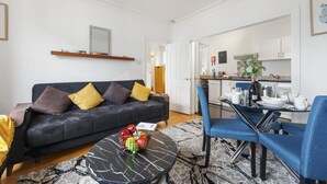 Ein modernes Wohnzimmer mit einer schwarzen Couch, einem Couchtisch mit Früchten und einem Essbereich mit blauen Stühlen und einem Glastisch.