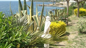 En statue af en figur i hvidt, omgivet af grønne planter og gule buske, med udsigt over et kystområde med bygninger og bakker i baggrunden.