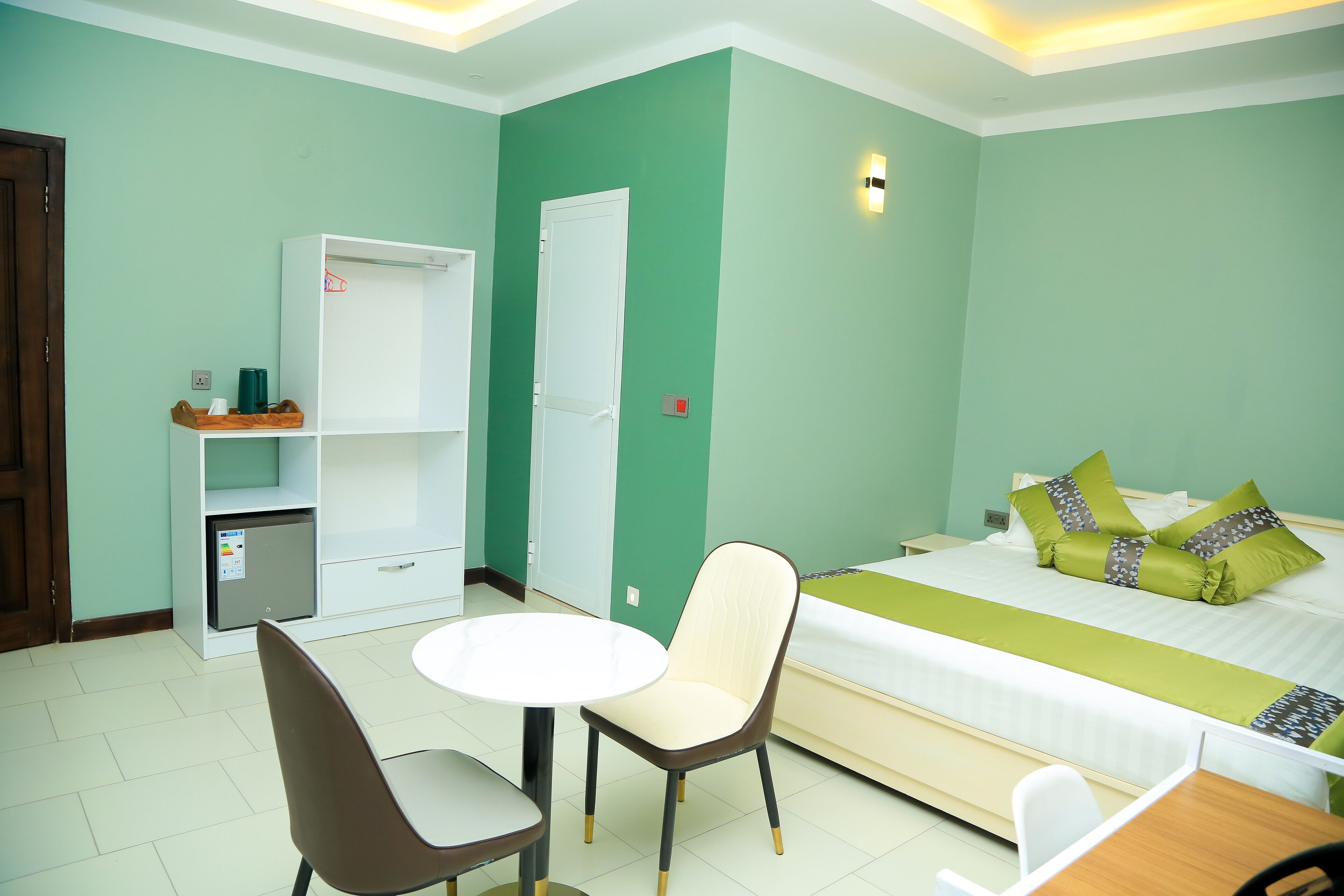 A hotel room with a bed, a desk, a chair, a small table, a wardrobe, and a mini-fridge.