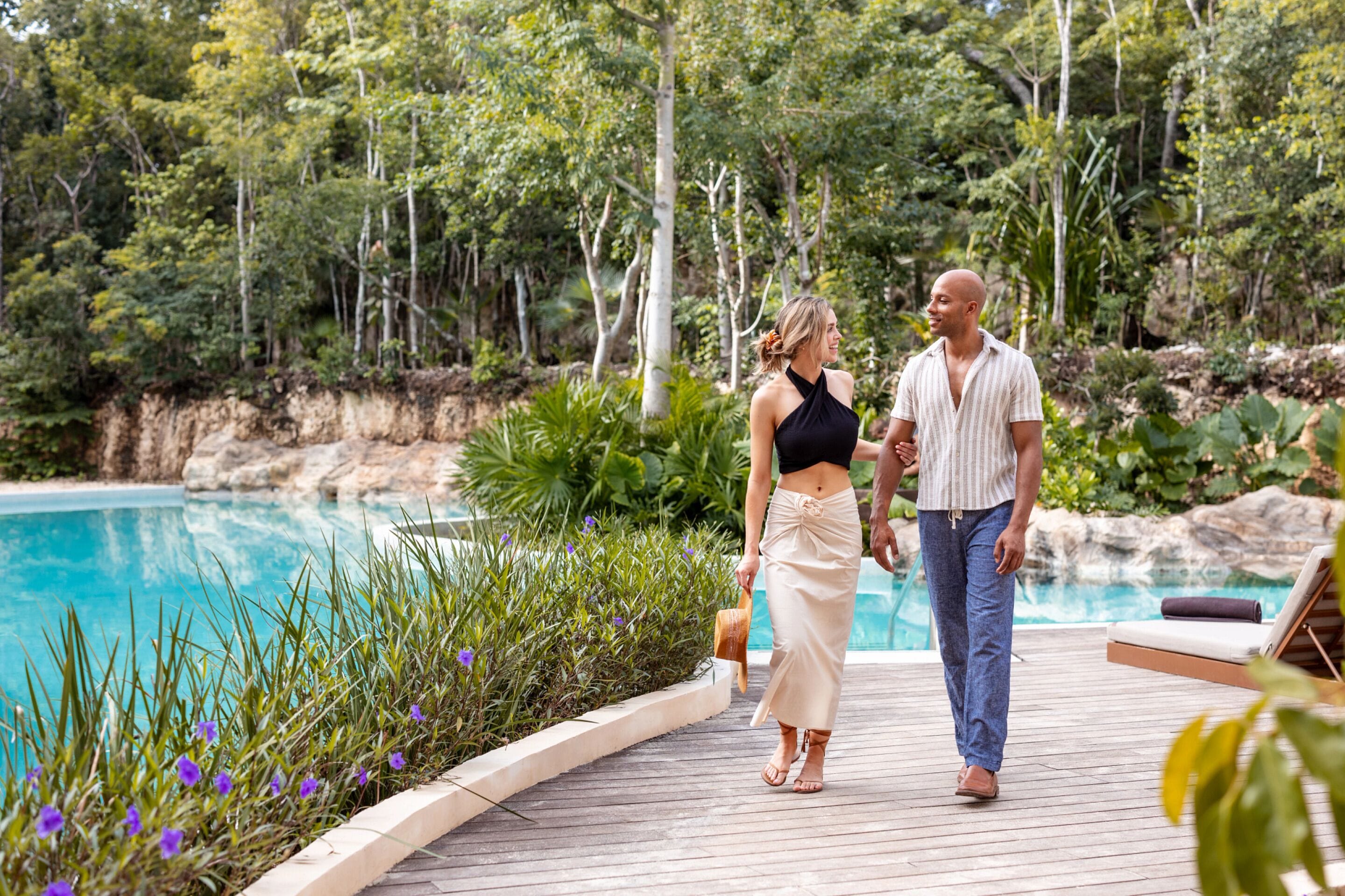 Una pareja caminando por una terraza de madera junto a una piscina, con exuberante vegetación y árboles al fondo.