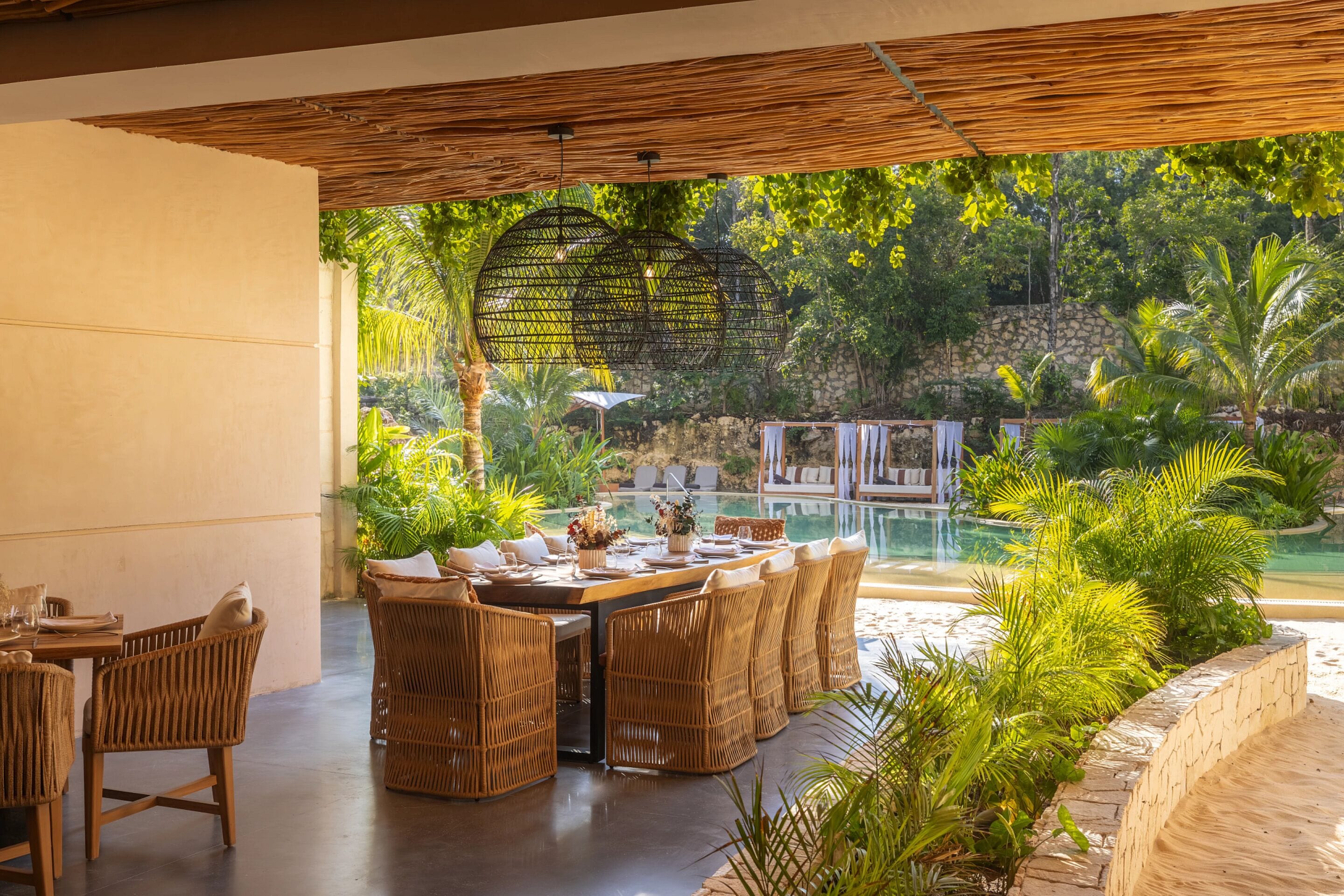 An outdoor dining area with wicker furniture and a view of a pool and lush greenery.