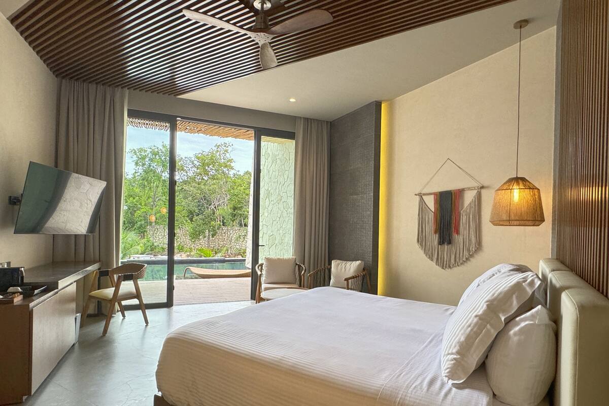 Un dormitorio moderno con una cama grande, un escritorio, una silla, un televisor en la pared y un balcón con vistas a zonas verdes.