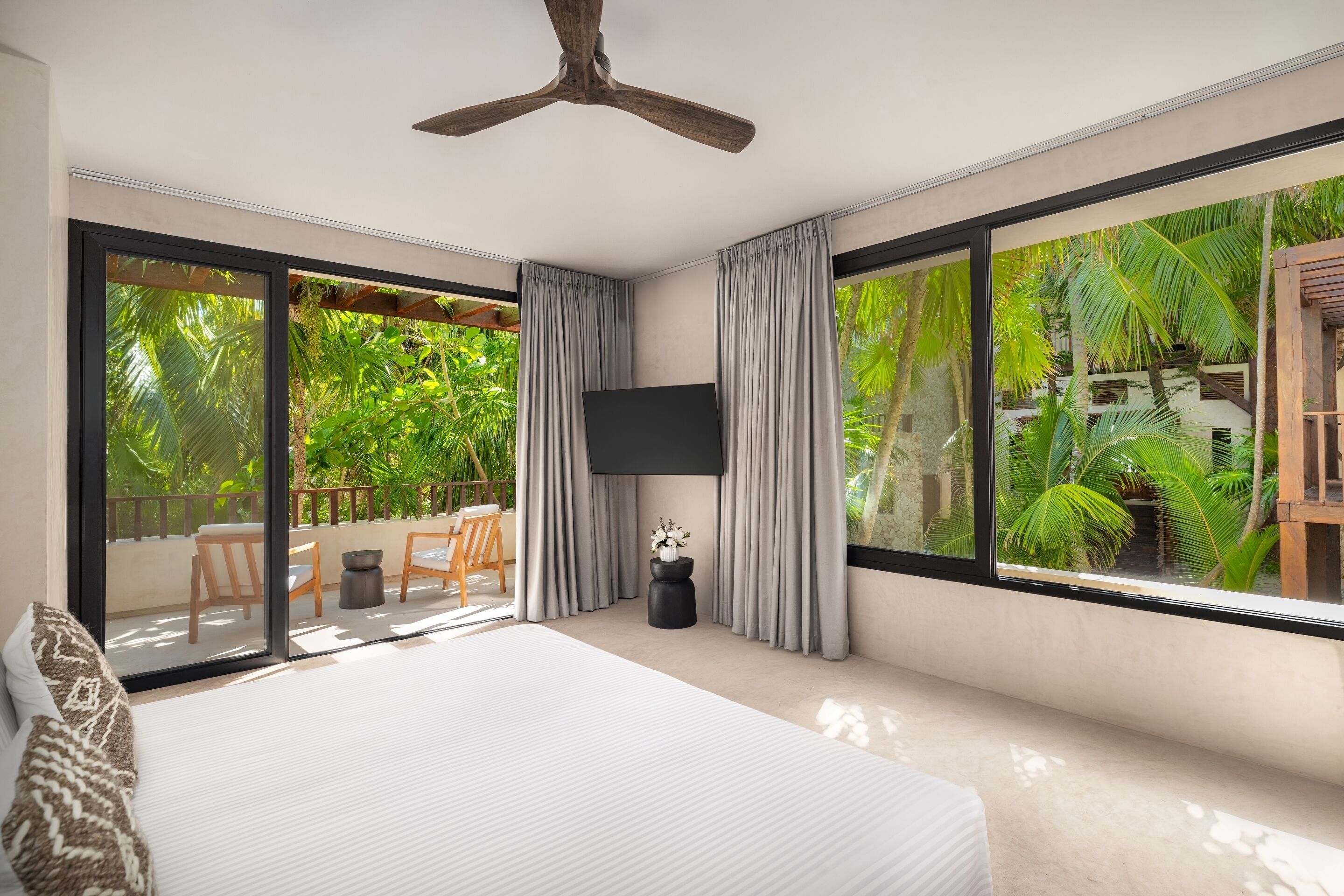 A modern bedroom with a large bed, a ceiling fan, a TV, and a sliding glass door leading to an outdoor area with a wooden deck and lush greenery.