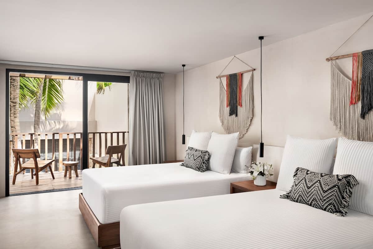 Una habitación de hotel moderna con dos camas, un balcón con sillas de madera y vistas a palmeras.