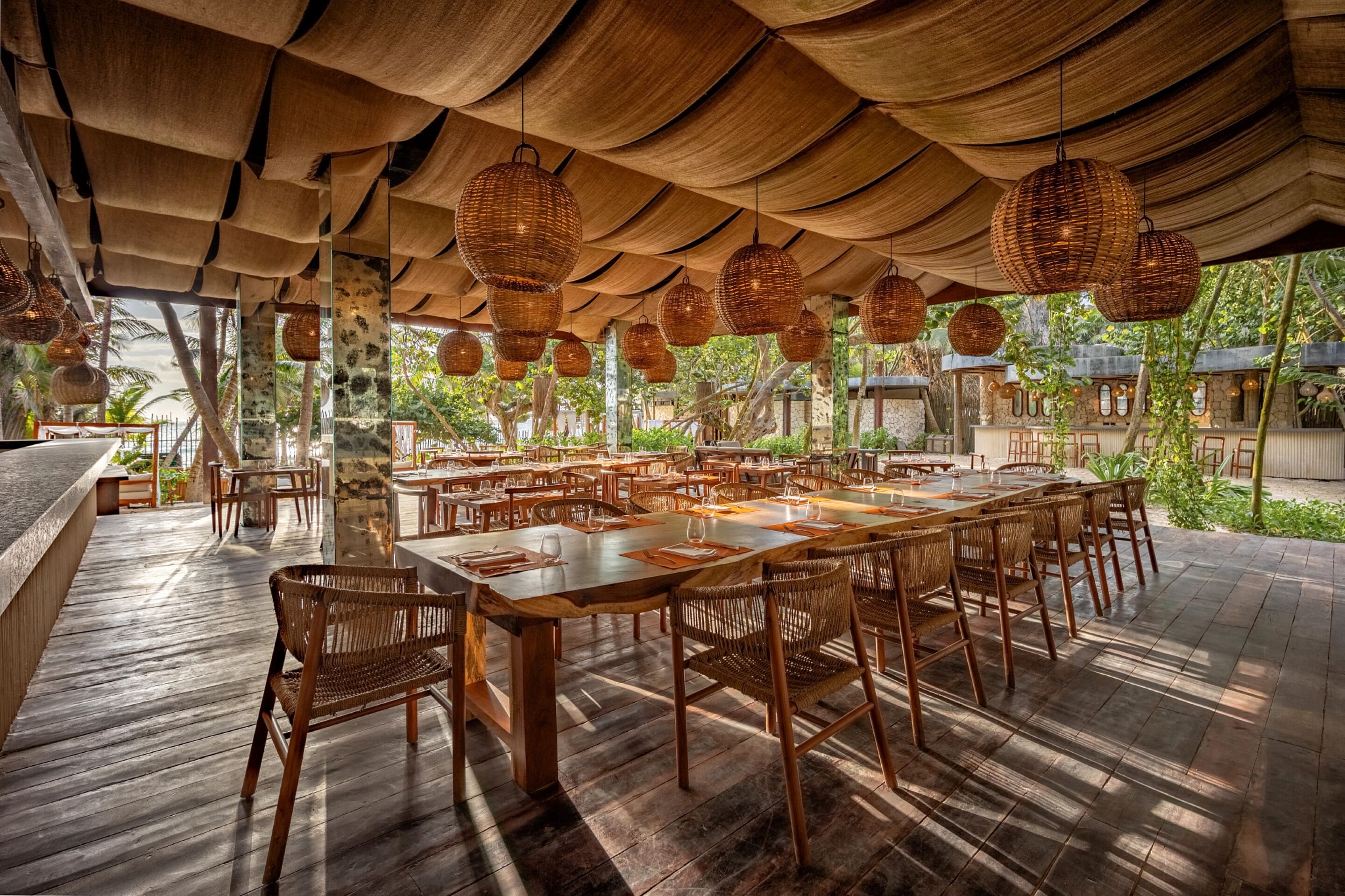An outdoor dining area with wooden tables and chairs, surrounded by a covered patio with woven ceiling lamps.