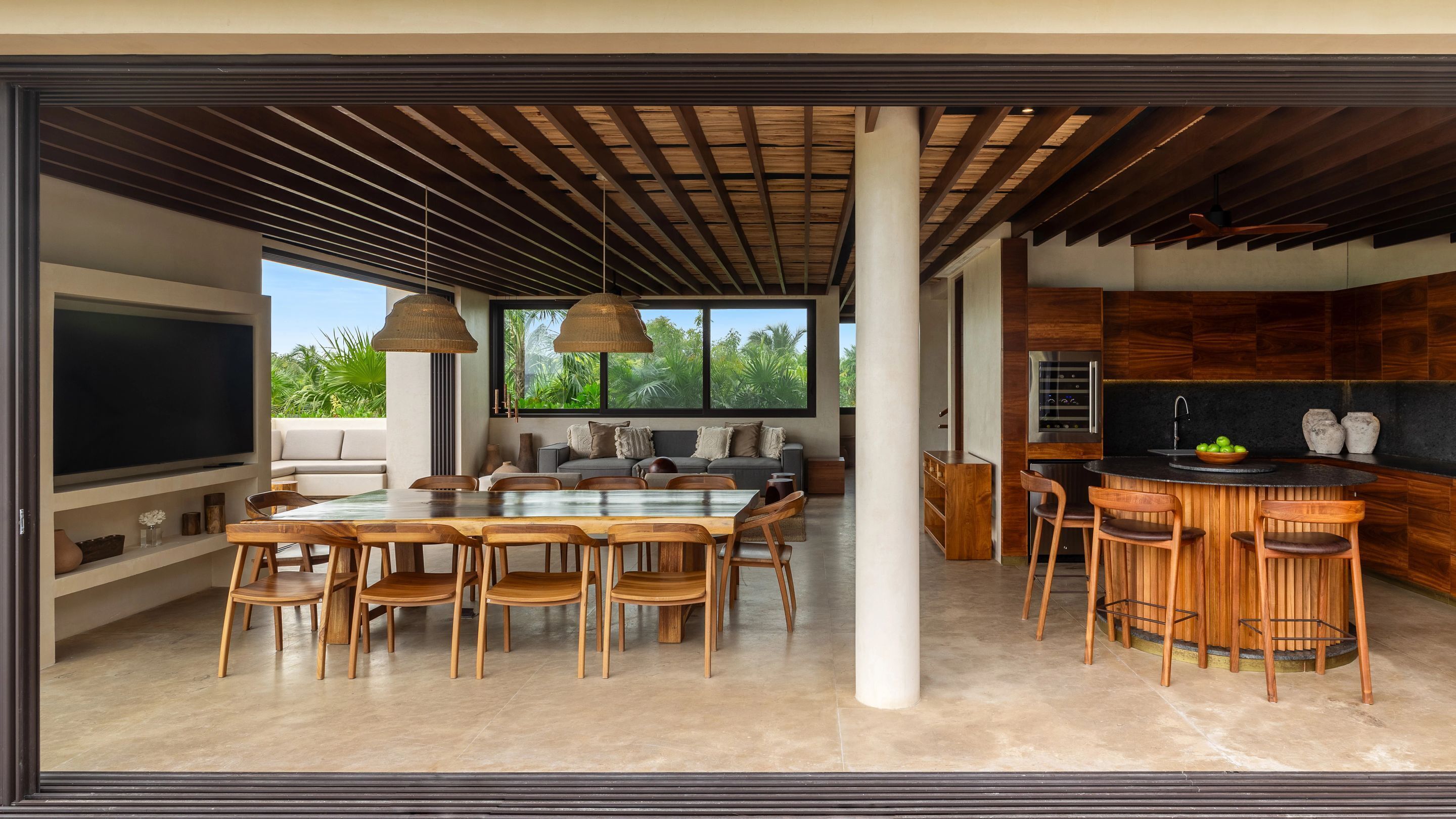 Ampla sala de estar com uma grande televisão, mesa de jantar e cadeiras, cozinha com armários de madeira e vista para o verde lá fora.