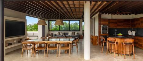 Ampla sala de estar com uma grande televisão, mesa de jantar e cadeiras, cozinha com armários de madeira e vista para o verde lá fora.