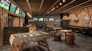 Een moderne barruimte met meerdere schermen waarop sportevenementen te zien zijn, een tafelvoetbaltafel en diverse zitmogelijkheden, waaronder ronde tafels en comfortabele fauteuils.