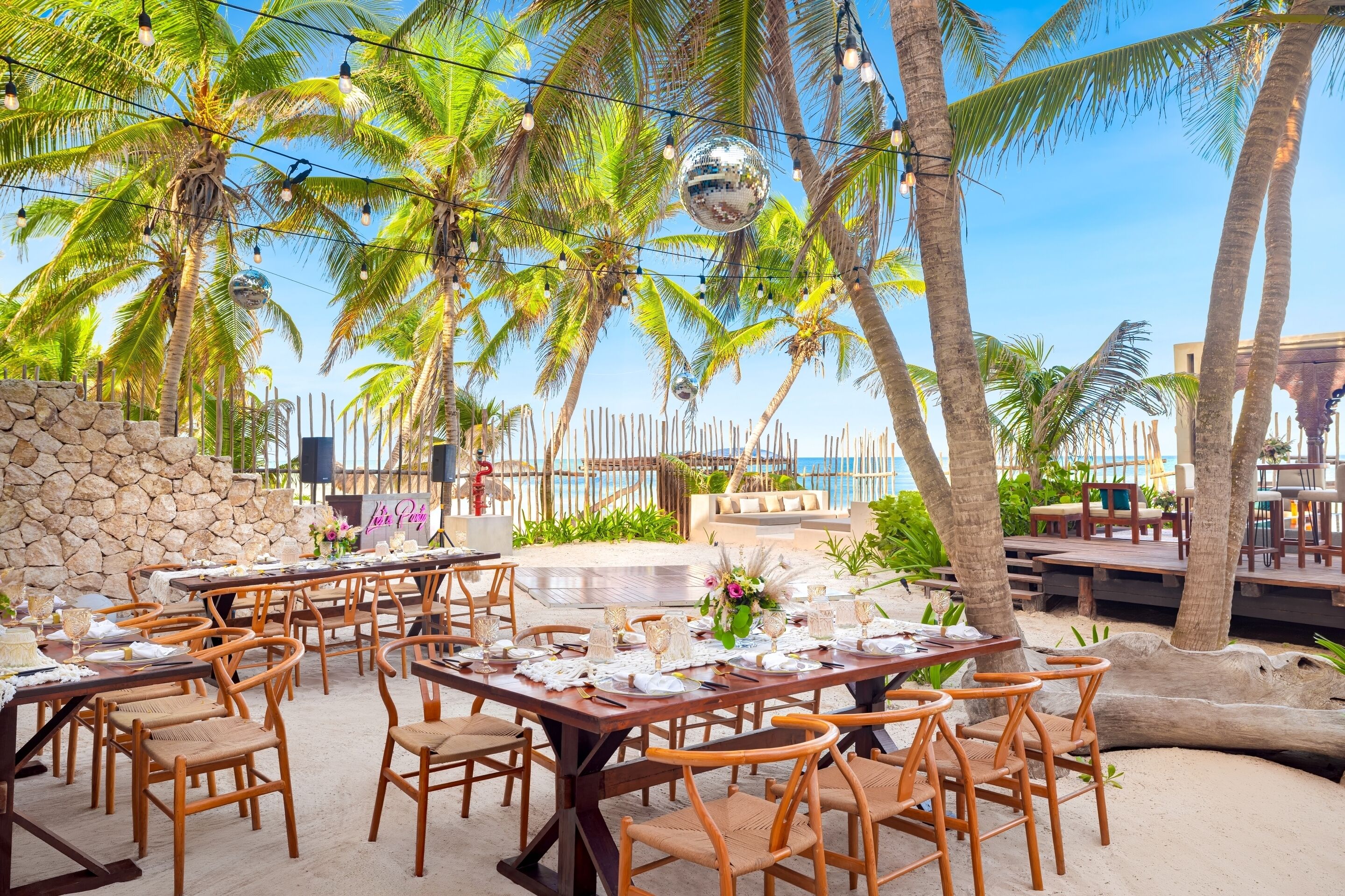 An outdoor dining area with wooden tables and chairs, palm trees, and a view of the ocean.