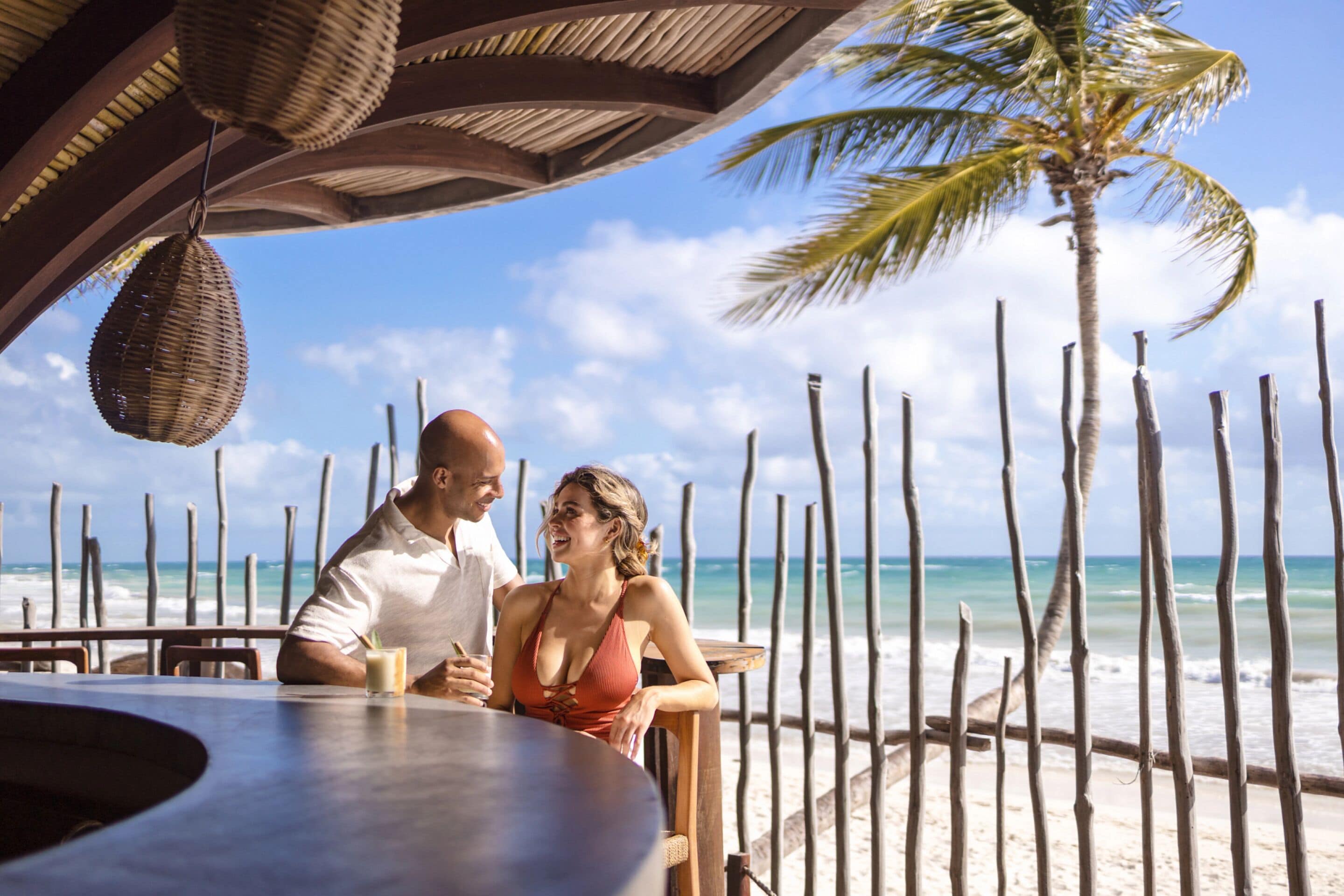 Una pareja cenando en un restaurante frente al mar, con vistas al océano y palmeras.