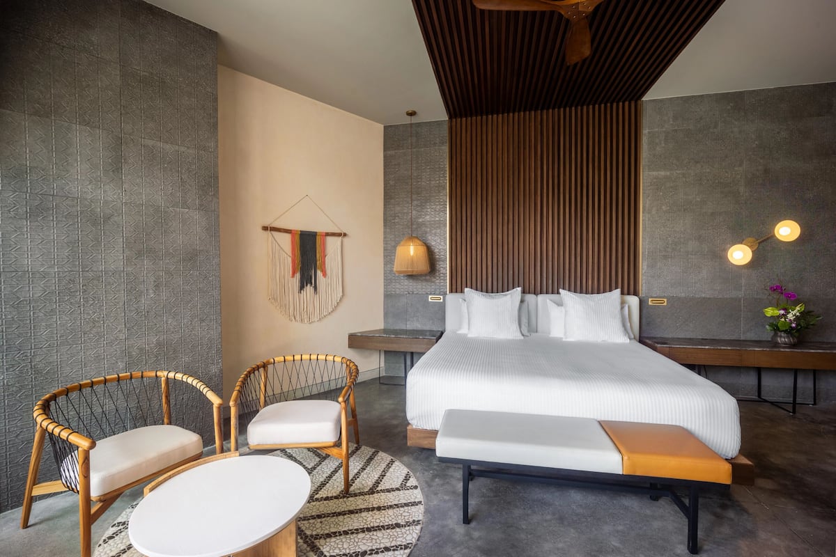 Habitación de hotel moderna con una cama grande, dos sillones de mimbre, una mesita redonda y un banco.