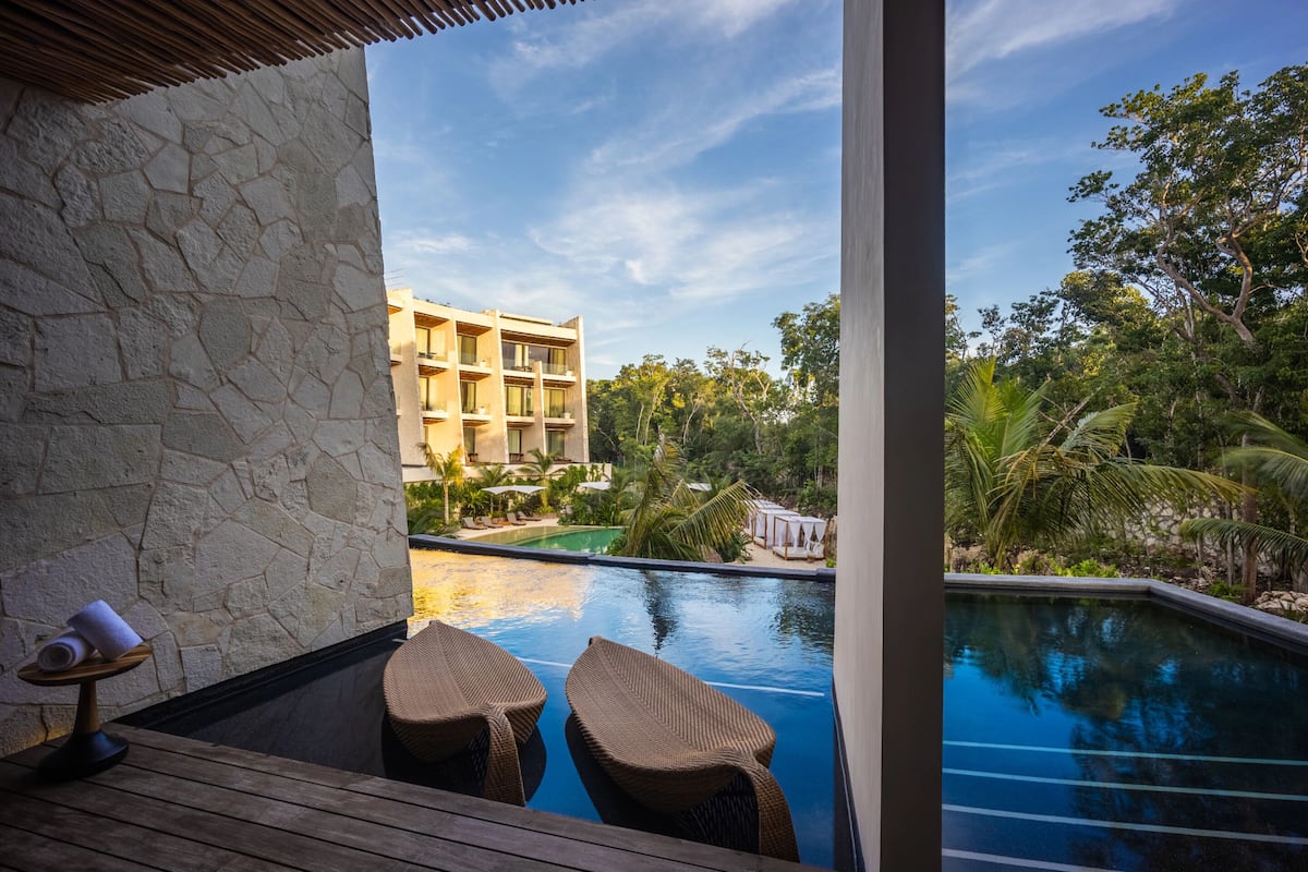 Zona junto a la piscina con sillas de mimbre, pared de piedra y vistas a un edificio y vegetación tropical.