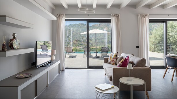 A modern living room with a television, sofa, and a view of a pool and mountains. - 3 Bedroom Modern Villa, Pool, Savoca, East Sicily (Savoca)