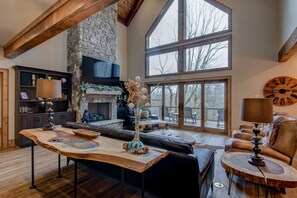 A spacious living room with a stone fireplace, large windows, and a high ceiling with exposed beams.