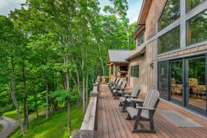 A wooden deck with outdoor seating and a stone fireplace, surrounded by lush greenery.
