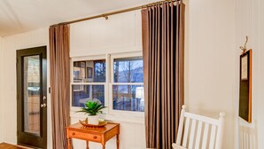 Ein Zimmer mit Holzboden, einem weißen Stuhl, einem kleinen Tisch mit einer Pflanze und einem Fenster mit Vorhängen.