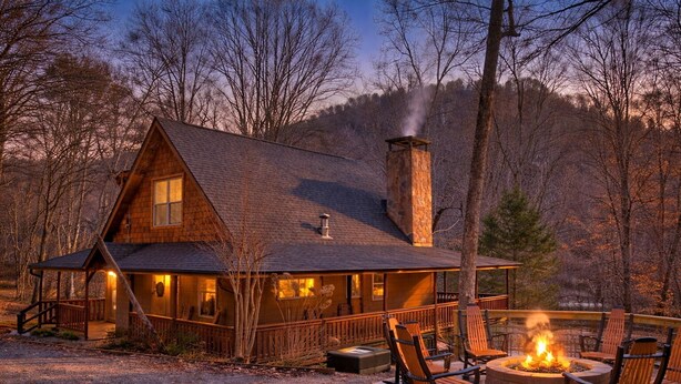 A log cabin with a stone chimney, a porch with chairs, and a fire pit.