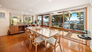A spacious dining area with a large wooden table and chairs, large windows with a view of the sea and boats, and a wooden deck outside. - Bancroft Haven: Nestled above the marina boasting views across Bancroft Bay (Metung)