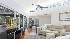 A modern living room with a flat-screen TV, a sofa, a ceiling fan, and large windows. - Karinyah - Panoramic Waterfront Views (Summerland Point)