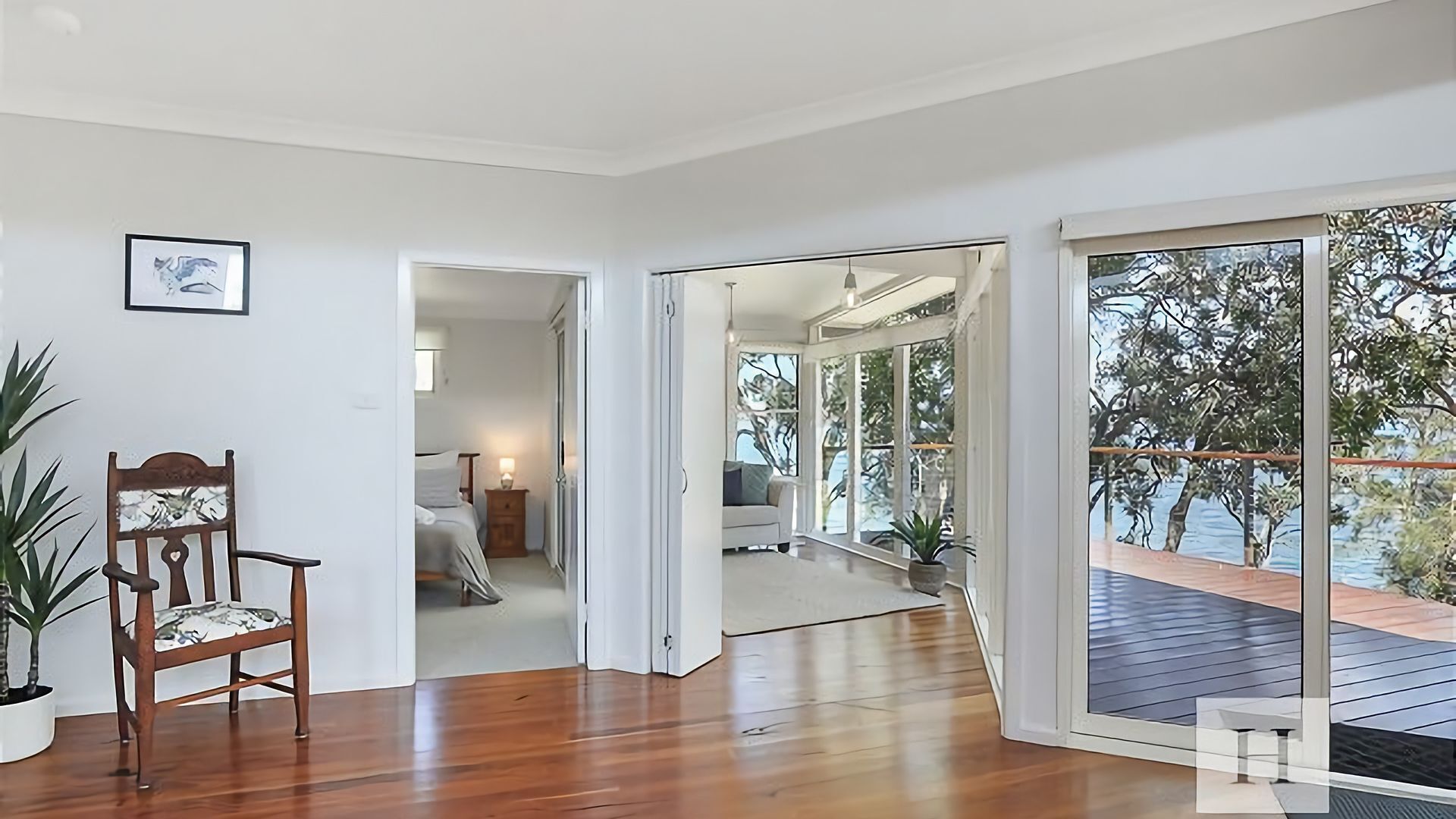 A spacious room with a wooden floor, a chair, a bed, and a large sliding glass door leading to a balcony with a view of trees.