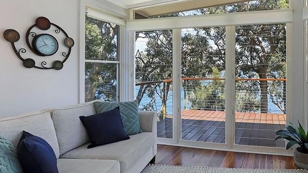 A modern living room with a sofa, a wall clock, and large glass doors leading to an outdoor deck. - Karinyah - Panoramic Waterfront Views (Summerland Point)