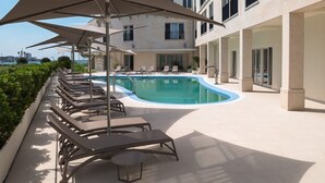 Zona de piscina con sillones de descanso y sombrillas, cielo despejado y un edificio con amplias ventanas.