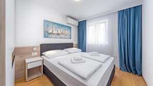 Una habitación de hotel moderna con una cama grande, una mesita de noche, un cuadro de veleros y cortinas azules.