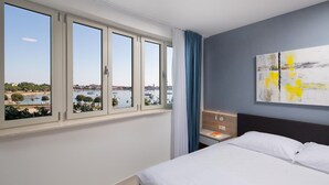 Ein Hotelzimmer mit einem Bett, einem Nachttisch, einem Gemälde und Blick auf ein Gewässer durch große Fenster.