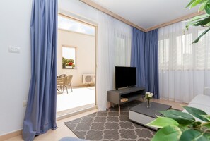 A modern living room with a flat-screen TV on a stand, a coffee table, and a sofa. There is a sliding glass door leading to an outdoor area with a chair and a potted plant.