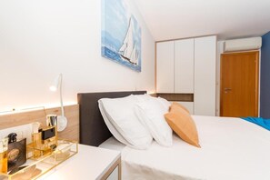 A hotel room with a bed, a bedside table with a lamp, a painting of sailboats, a wardrobe, and an air conditioning unit.