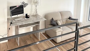 Ein modernes Wohnzimmer mit Holzboden, einem weißen Sofa, einem Glastisch, einem Schreibtisch mit Computer und einer Lampe.