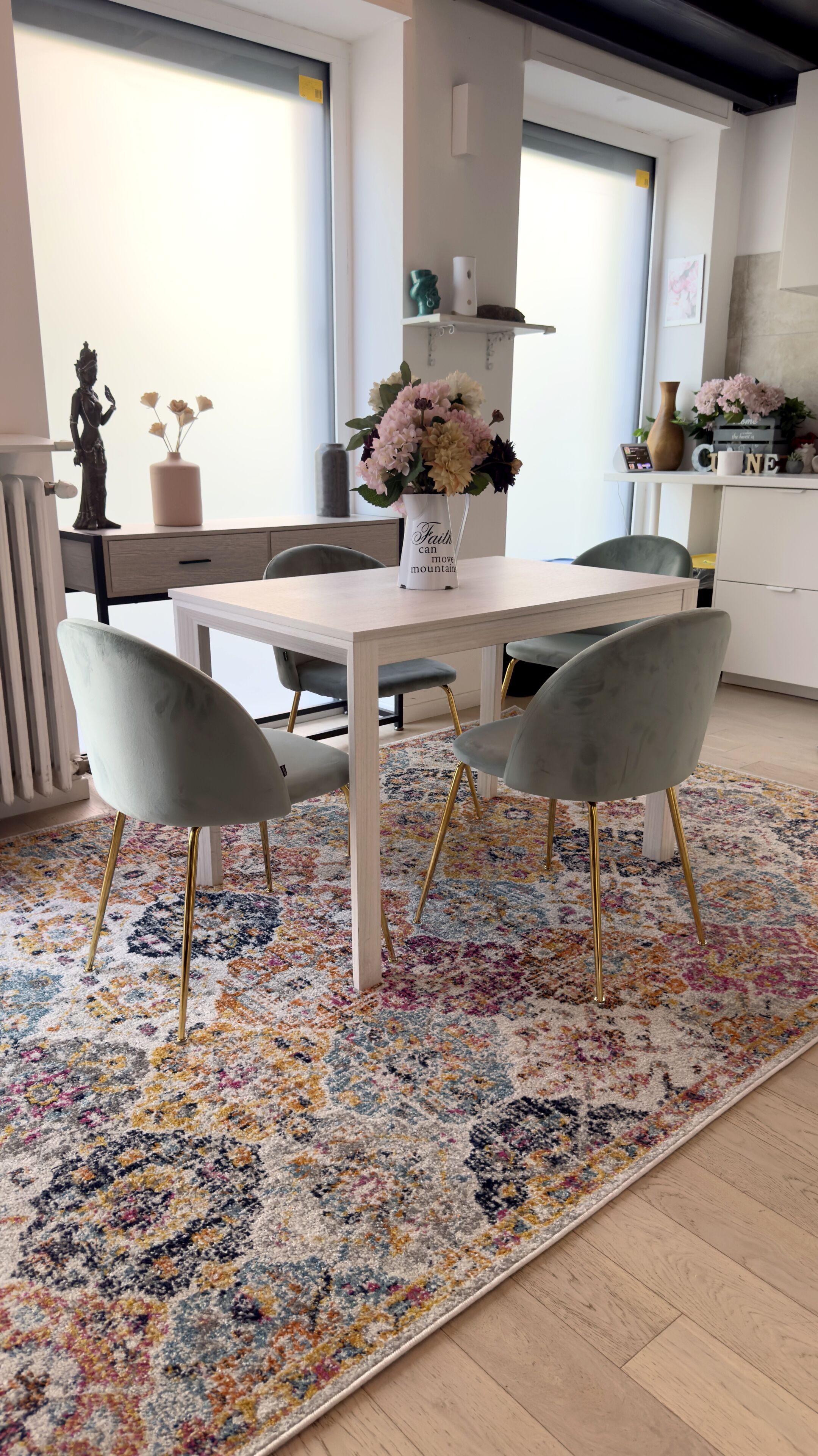 Un coin repas avec une table blanche, deux chaises grises, un tapis à motifs floraux et une statue sur la gauche.