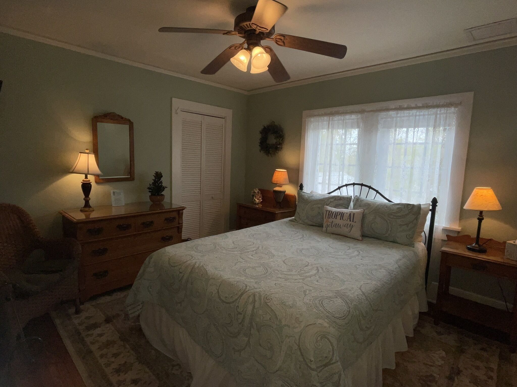 Une chambre à coucher comprenant un lit, un ventilateur de plafond, une fenêtre avec des rideaux, une commode et deux lampes de chevet.