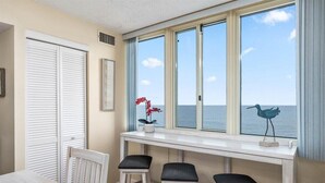 Une pièce avec vue sur la mer, une table blanche avec deux chaises et une fenêtre ornée d’une sculpture d’oiseau.