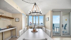 Un espace repas avec une grande table, des chaises blanches, un lustre et une vue sur l’océan grâce à de grandes fenêtres.