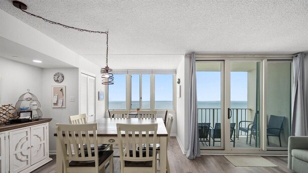 Un comedor con una mesa y sillas, un reloj en la pared y vistas al mar a través de grandes ventanales.