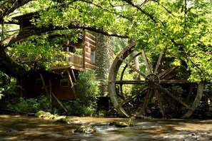 A rustic wooden structure with a large water wheel, surrounded by lush greenery and a flowing stream. - Lindsey Mill Cabin on stream and pond (Gatlinburg)