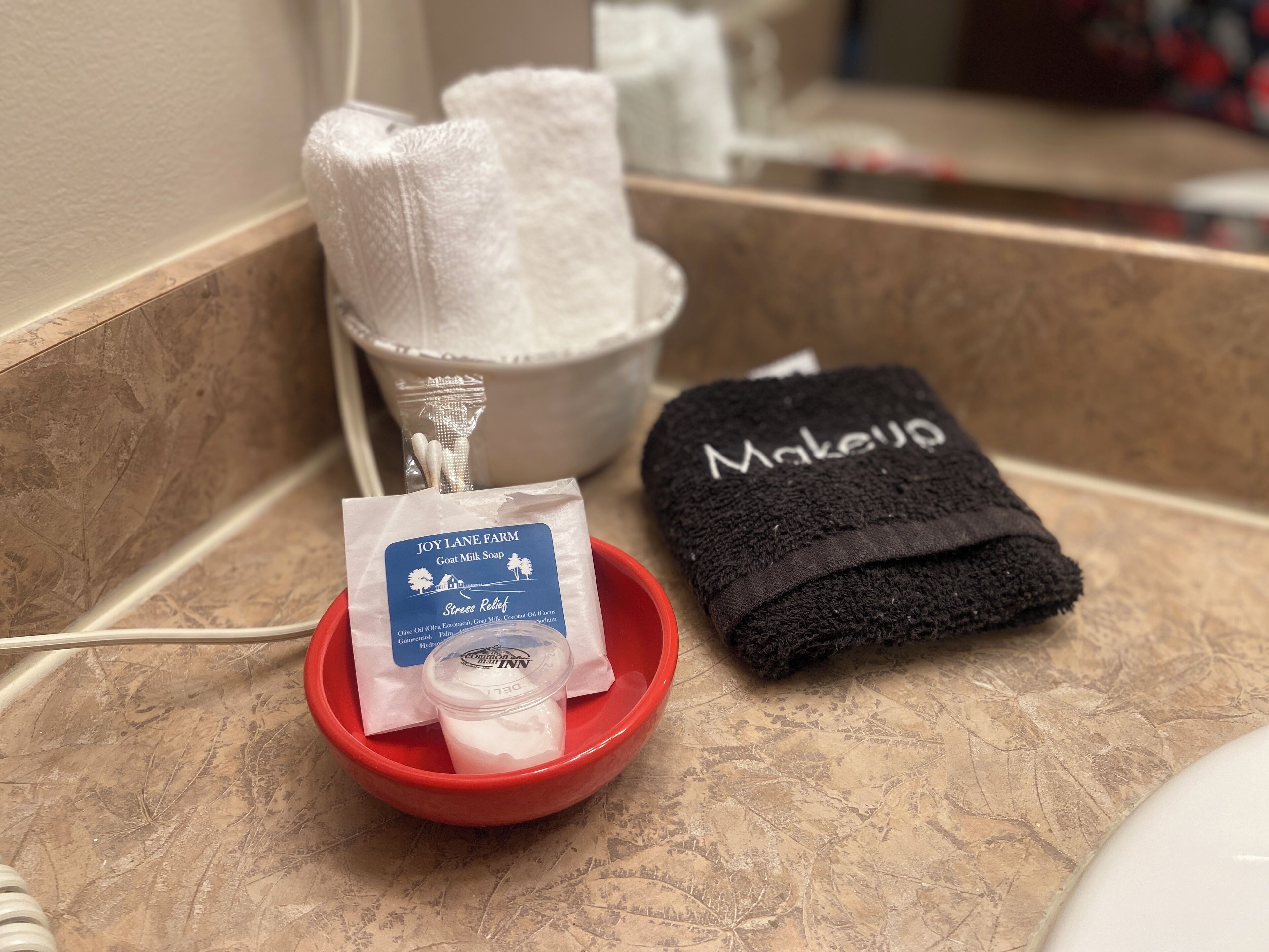 Un comptoir de salle de bain avec un bol rouge contenant un paquet de savon au lait de chèvre, une serviette noire sur laquelle est écrit le mot « Maquillage », et une serviette blanche dans un récipient blanc.