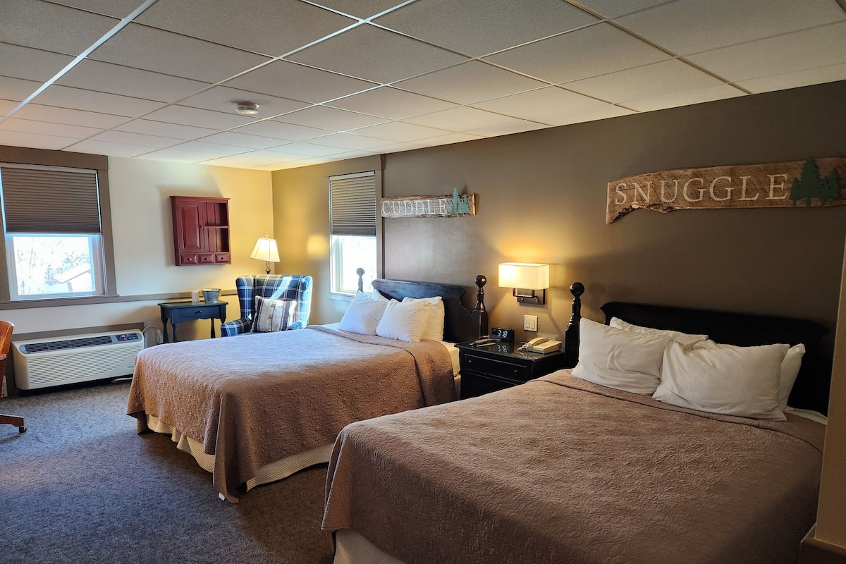 A hotel room with two beds, a desk, a chair, a lamp, and a thermostat.