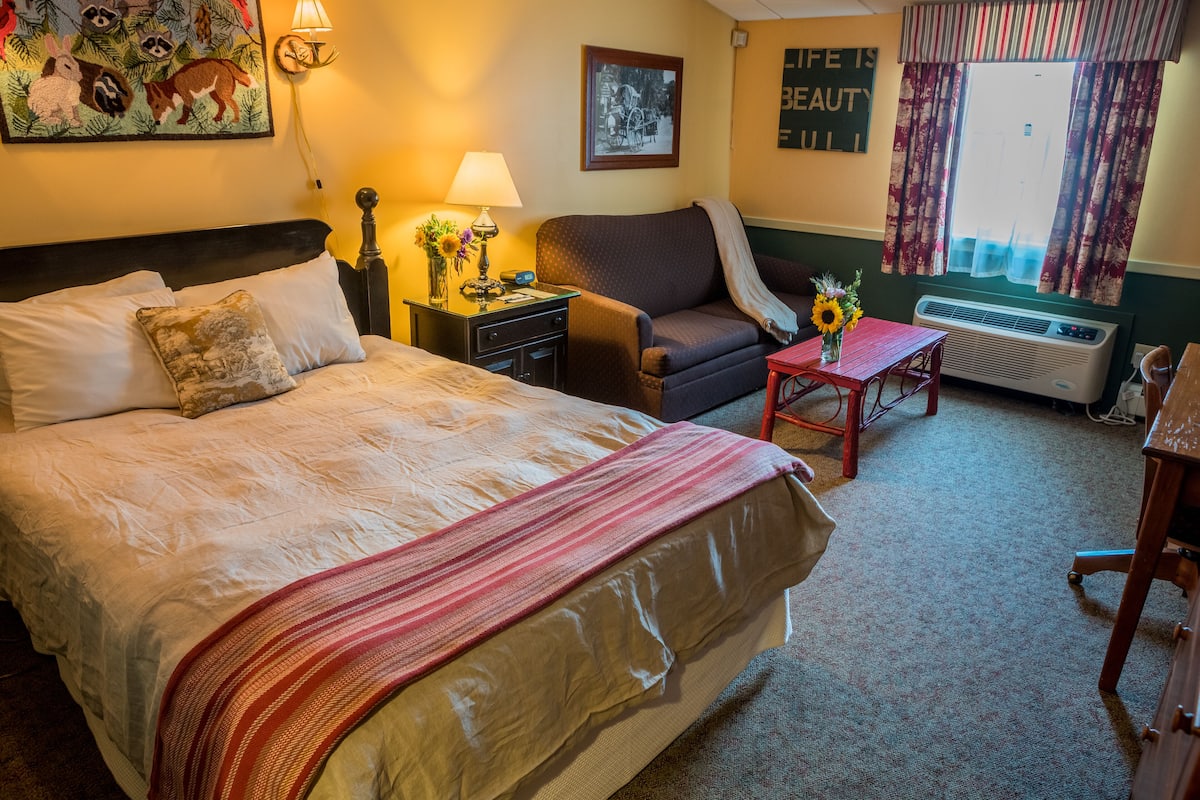 A hotel room with a large bed, a sofa, a small table with a vase, a lamp, and a picture on the wall.