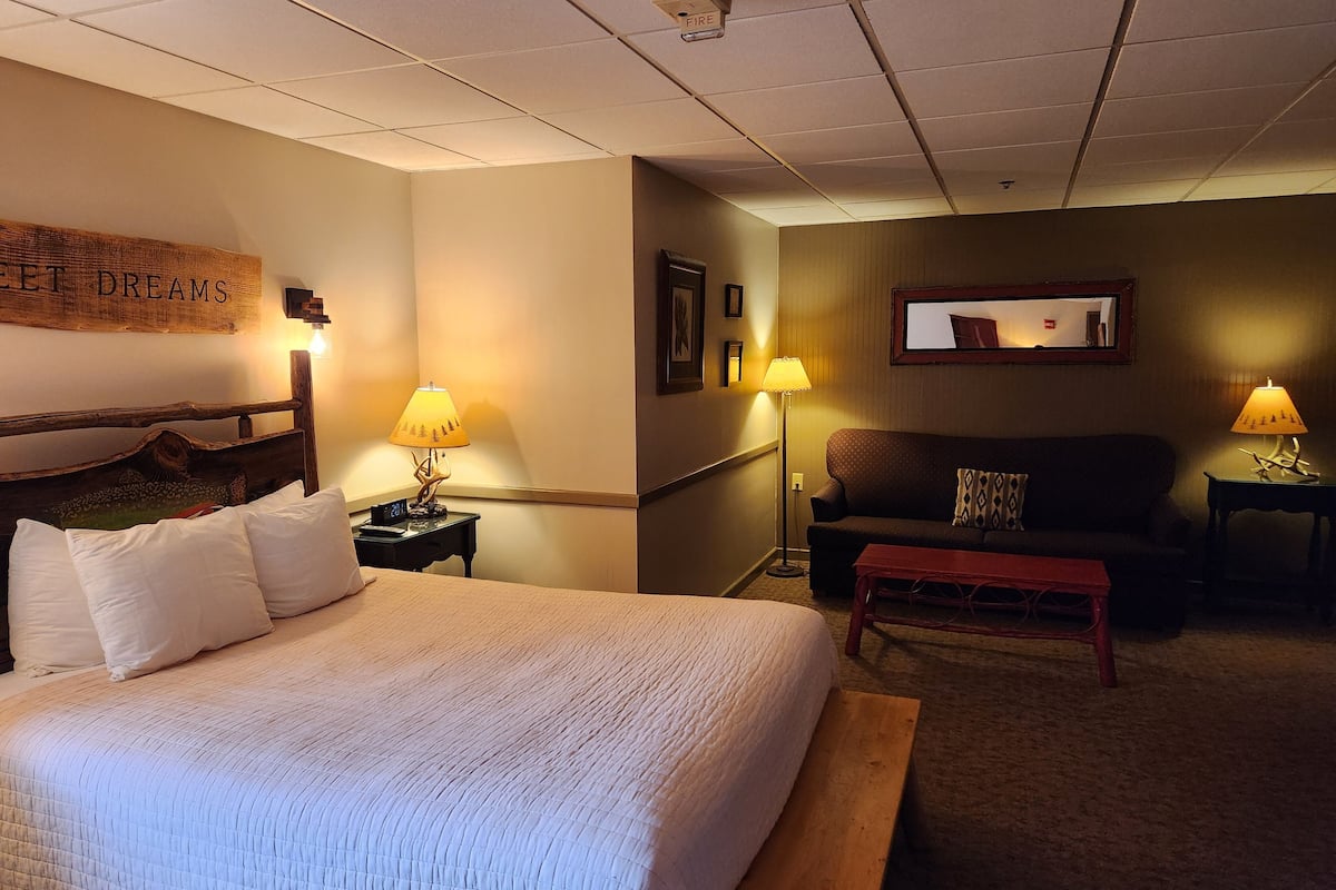 A hotel room with a large bed, a wooden headboard, two bedside tables with lamps, a sofa, a small wooden bench, and a mirror on the wall.