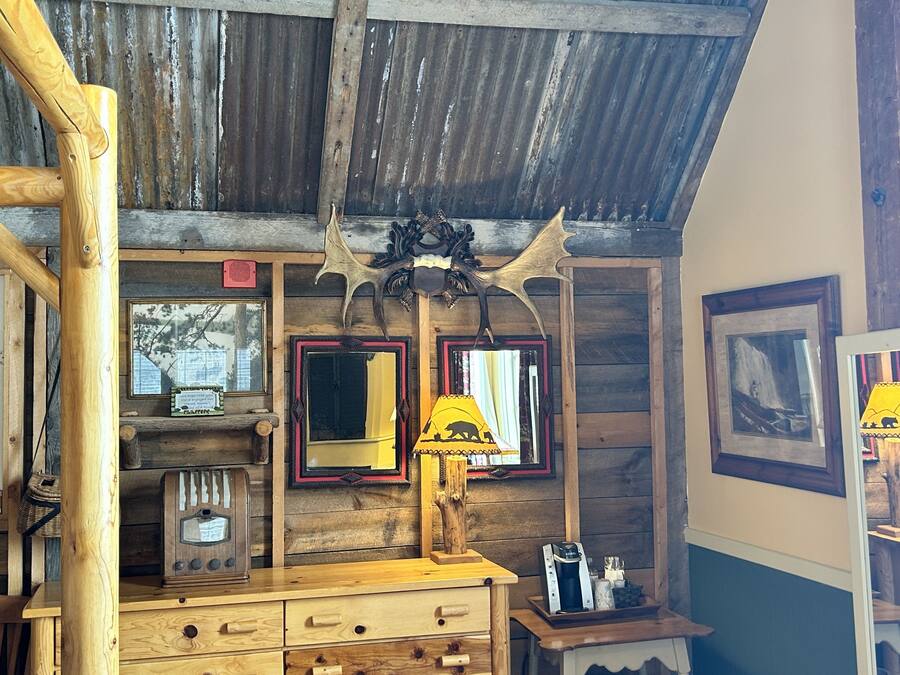 A rustic cabin interior with wooden walls, a wooden dresser, a lamp, and framed pictures.