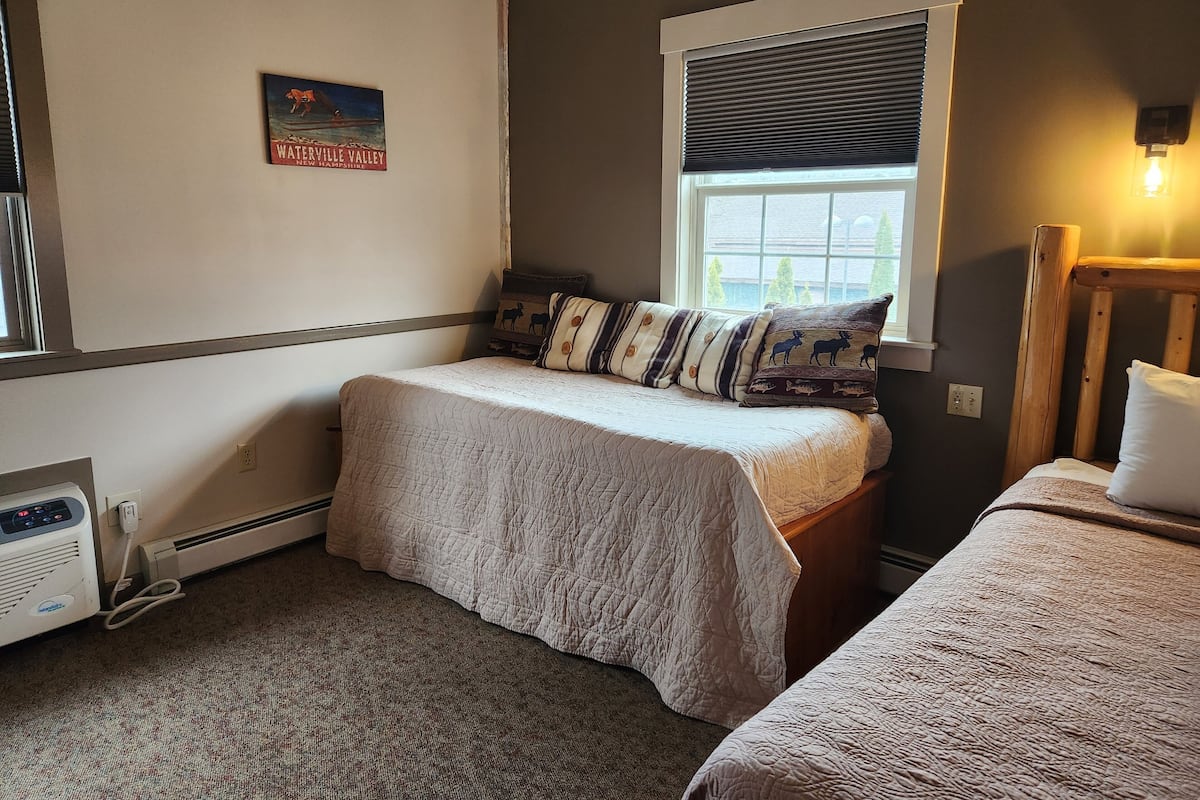 A bedroom with two beds, a window with blinds, a heater, and a wall-mounted light fixture.
